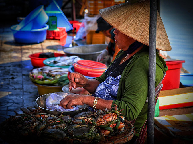 Excursions while on a Mekong Delta Cruise A woman selling fish from a bowl, sitting on the floor, wearing a traditional Vietnamese hat