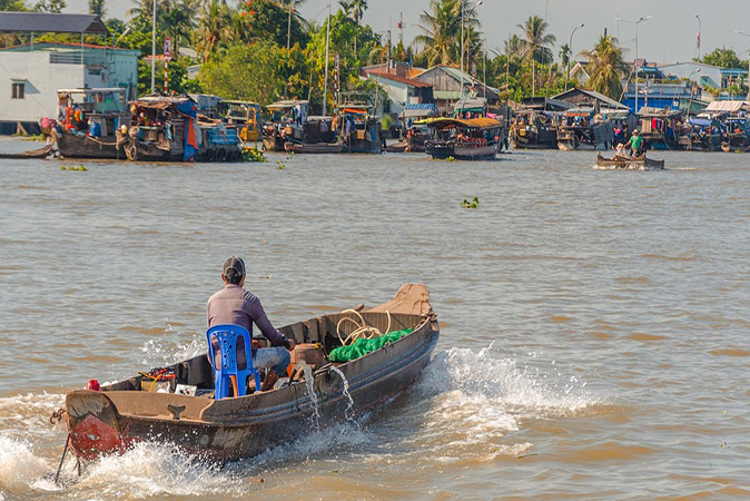 the best time for a Mekong River cruise A man sitting on a blue plastic chair inside a very basic wooden boat, heading towards shore where there are many more boats, small houses and greenery