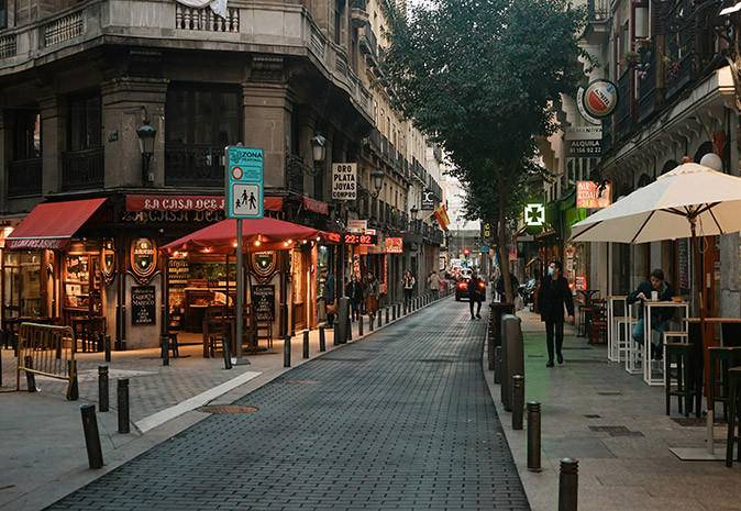 Things to know about safety and crime in Madrid A typical Spanish street after dark with cafes, several pedestrians, trees, and apartment buildings lining both sides of the street