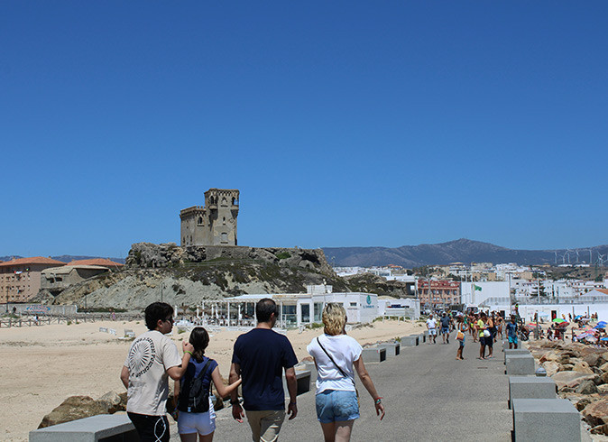 Life in Tarifa, Spain Various people on a boardwalk with various buildings and ruins of a castle in the background