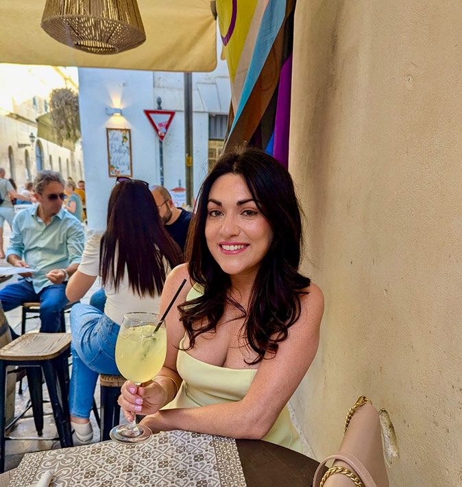 A woman sitting on a terrace, holding a large yellow looking cocktail in a wine glass, smiling at the camera