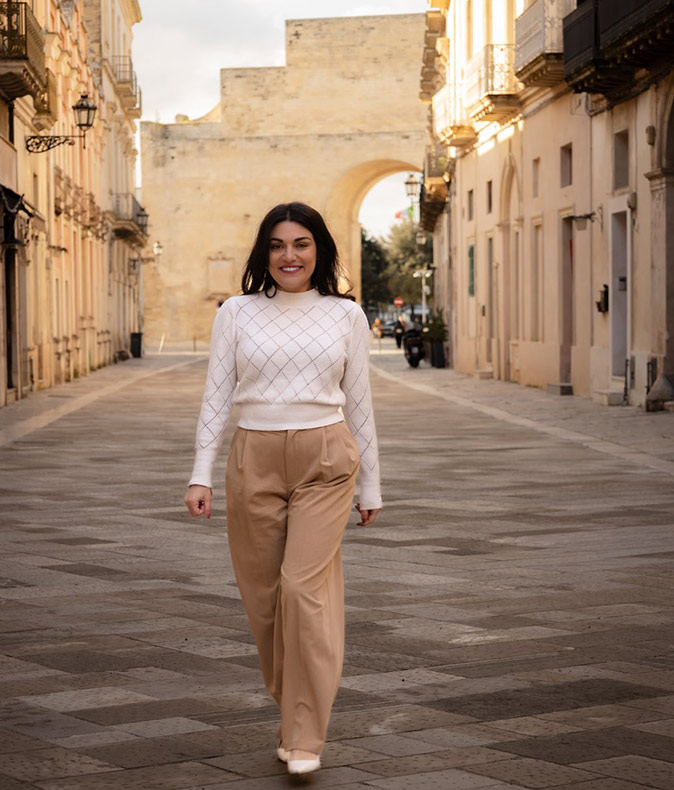 A woman walking towards the camera, smiling, surrounded by typical Italian buildings in beige and brown