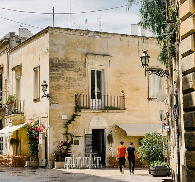 2 people walking in front of a large, beige building that shows 2 restaurant terraces and some plants