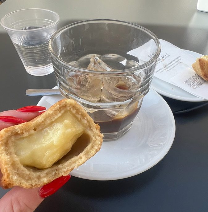 Part of a hand holding a cream filled pastry with a coffee cup showing on the table behind the hand