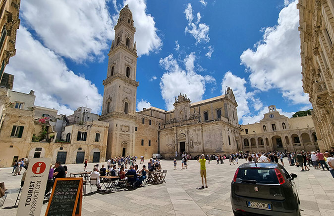 A large Italian city square showing a church, several other beige and brown buildings, a terrace filled with people, several pedestrians and a parked car