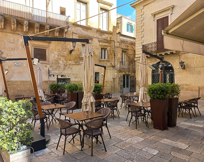 An empty restaurant terrace with brown tables and chairs, surrounded by beige and brown buildins and some plants