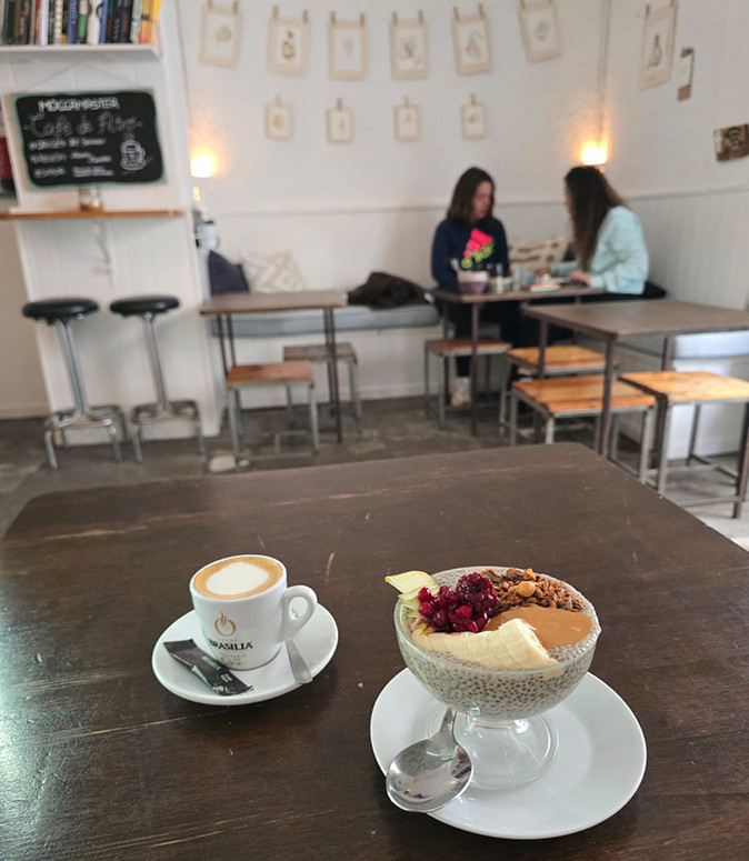 Working remotely from Tarifa, southern Spain A coffee and a breakfast bowl on a wooden table with other tables and 2 cafe guests in the background