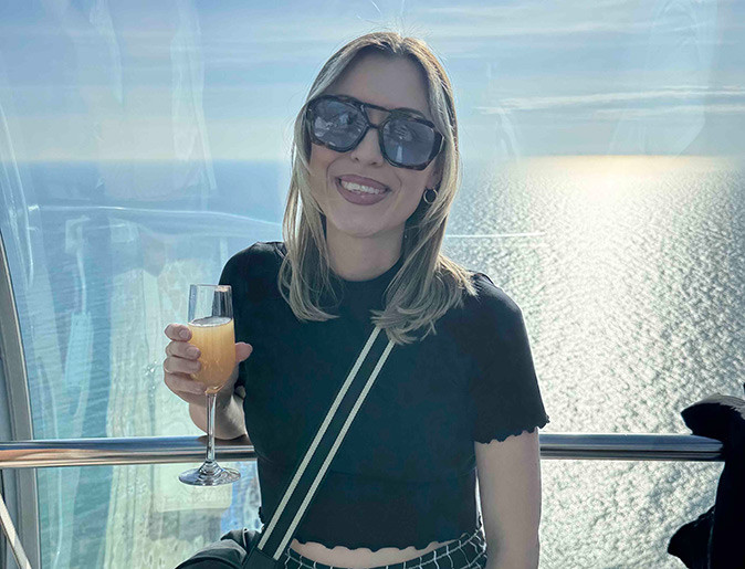 A woman wearing sunglasses and holding a drink, smiling at the camera, with the sea in the background