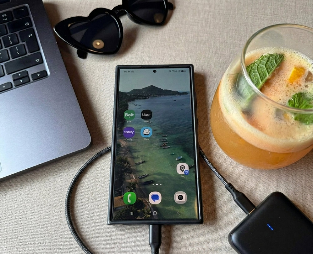 A phone showing various downloaded apps, plus part of a laptop, sunglasses, a powerbank and an orange drink