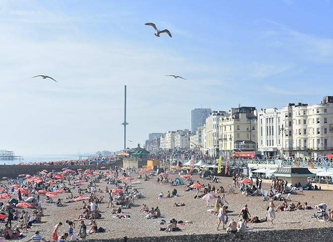 A large sandy beach filled with people, some seagulls flying, and several white and gray buildings in the background