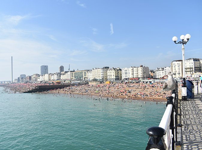 Photo taken from a pier overlooking a sandy beach filled with people, with several white and gray buildings in the background