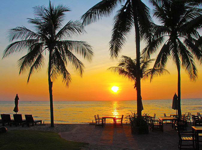 Visiting Hua Hin, Thailand A warm, orange sunset over a sandy beach with palm trees and some tables and chairs