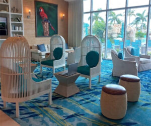 A hotel lobby with various white and blue chairs, small tables and a laptop on one of the tables. Palm trees outside a large window suggest a tropical hotel location