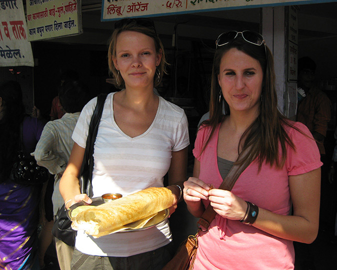 2 women standing in front of Indian signs holding an Indian dhosa on a plate