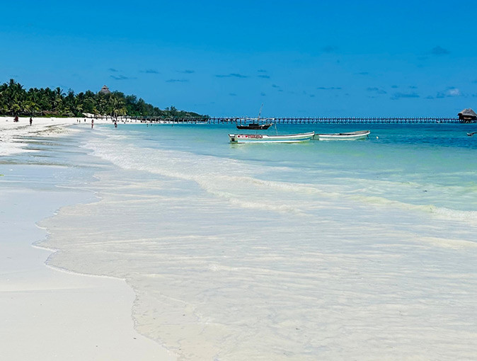 A white sandy beach with palm trees, some boats and a pier in the background