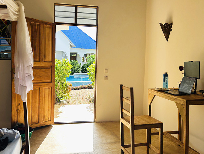A room with a wooden desk and a laptop, with the door open showing the pool outside