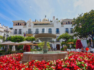 An urban setting with a fountain surrounded by red flowers with a white, historic Spanish building in the background