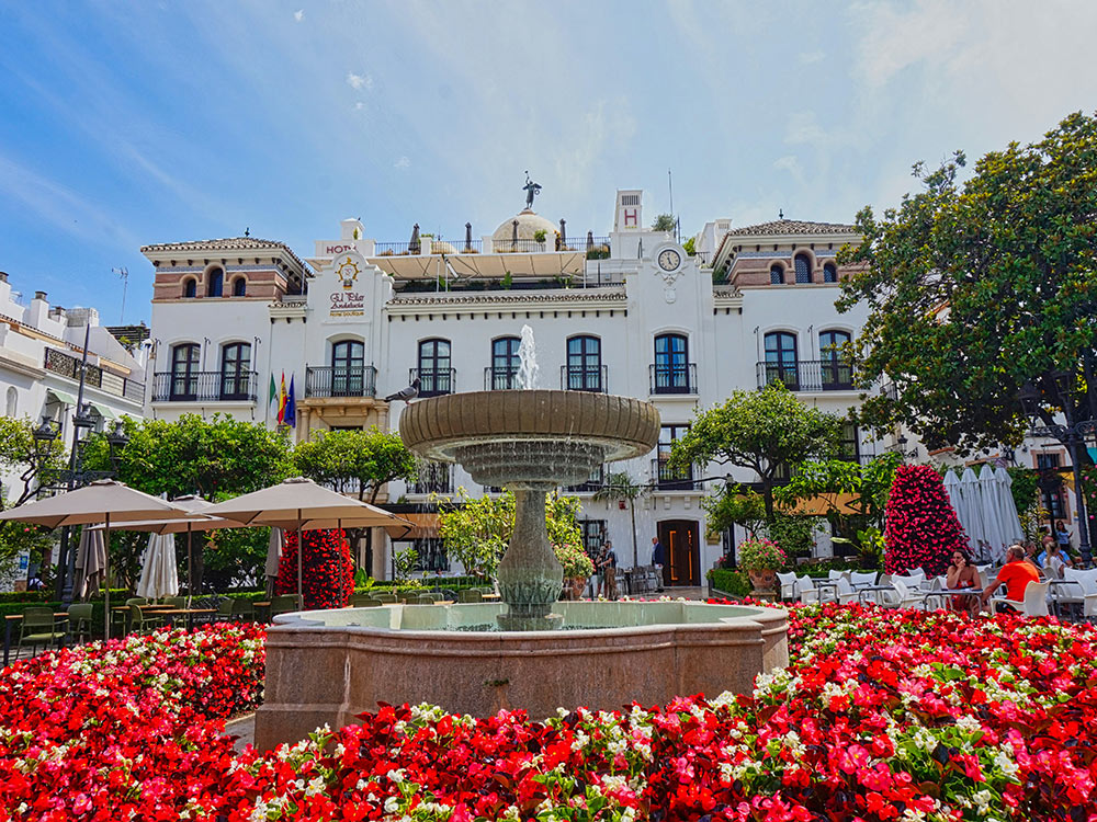 An urban setting with a fountain surrounded by red flowers with a white, historic Spanish building in the background