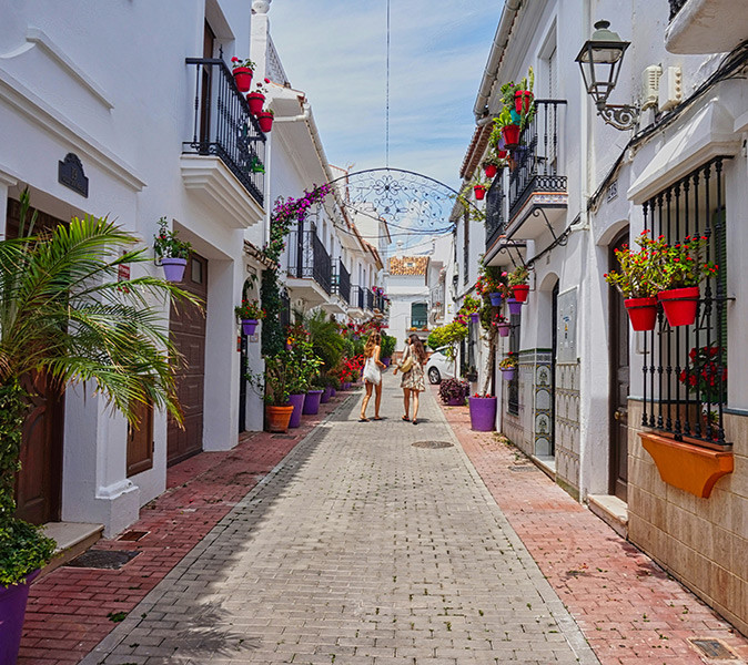 A pedestrianized street with whitewashed houses, many flower pots and 2 women walking