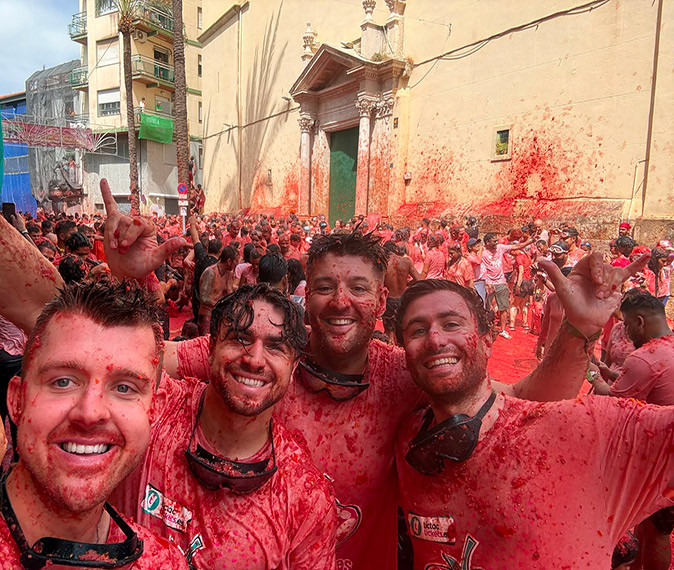 The biggest tomato fight 4 men smiling at the camera, covered in tomato pulp, in a crowd of people all covered after the tomato fight
