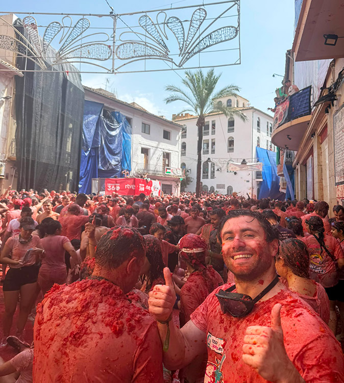 reviewing La Tomatina festival in Spain A man covered in tomato pulp giving a thumbs up, in a crowd of people covered in tomato pulp at the end of the La Tomatina event